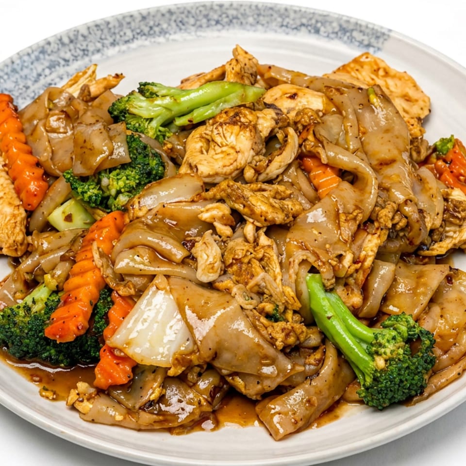 Plate of stir-fried wide noodles with chicken, broccoli, carrots, onions, and crispy toppings.